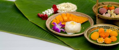 Pirinç tepside servis edilen geleneksel Tayland tatlıları ve yasemin çelengi ve beyaz masa üzerine süslenmiş muz yaprakları ile ahşap tepsi yakın görünüm