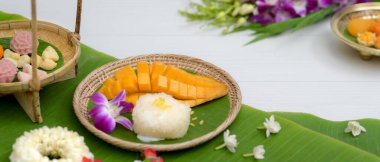 Tahta tepside servis edilen çeşitli Tayland geleneksel tatlıları. Beyaz masada çiçek ve muz yapraklarıyla süslenmiş.