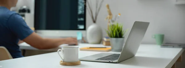 Side view of portable workspace with laptop, cup, stationery and decorations on white desk in blurred office room background