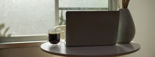 Portable workspace on white coffee table with open laptop, coffee mug and flower vase next to window