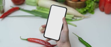 Mutfak masasında taze sebzelerle yemek pişirmek için gerekli malzemeyi hazırlarken akıllı telefondan kadın tarifini yakından izle. 