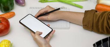 Bir kadının boş defterden yemek tarifi notları alırken çakma akıllı telefondaki bilgilere bakarken kenar görünümü 