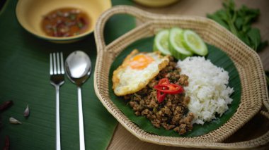 Kırpılmış domuz pirzolası, fesleğenli (Pad ka prao), hasır tabakta servis edilen pirinç sahanda yumurta ve gümüş çatal bıçak.
