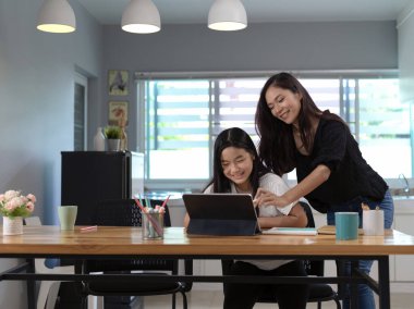 Oturma odasındaki ahşap tabletle ilgili ödev yapmasına yardım eden annenin portresi. 