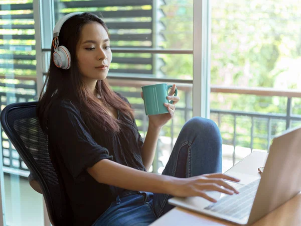 Side view of female relaxed woman with headphone using laptop and holding a coffee cup while sitting at worktable in bed room