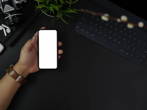 Overhead shot of hand holding blank screen smartphone above dark modern office desk with office supplies and decorations