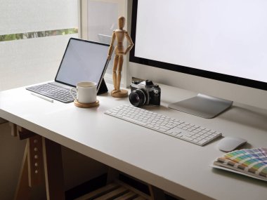 Model bilgisayar, dijital tablet, kamera, tasarımcı malzemeleri ve pencere kenarındaki dekorasyonla rahat ofis masasının kırpılmış görüntüsü. 