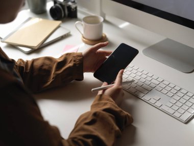 Beyaz ofis masasındaki akıllı telefondan bilgi ararken bilgisayar aygıtıyla çalışan bir kadının görüntüsü. 