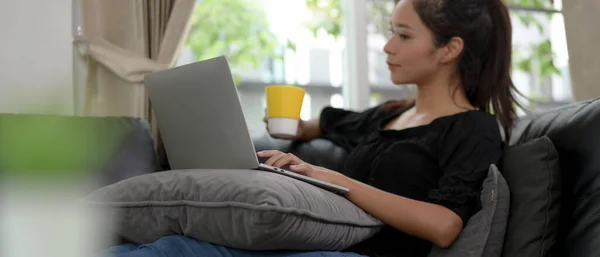 Close up view of young female lying relaxed on sofa and holding a cup of coffee while using laptop