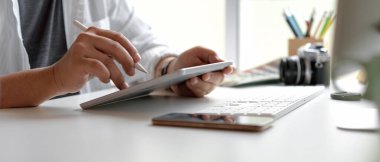 Beyaz ofis masasında akıllı telefon, bilgisayar aygıtı ve diğer malzemelerle stilus kalemle dijital tablet kullanan erkek tasarımcının yan görünümü 