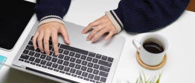 Beyaz ofis masasında, tablet, kahve fincanı ve ağaç kabıyla dizüstü bilgisayar kullanan kadın çalışanın kırpılmış görüntüsü. 