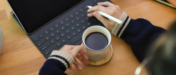 Top view of female left hand holding coffee cup and right hand using digital tablet above worktable