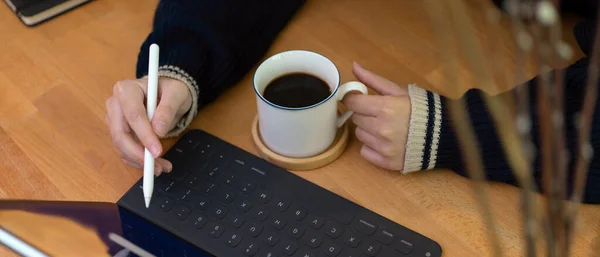 Close up view of female left hand holding coffee cup and right hand using digital tablet above worktable