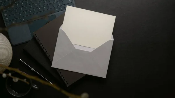 Open mock-up invitation card with grey envelope above black schedule books on dark modern office desk with supplies and decoration