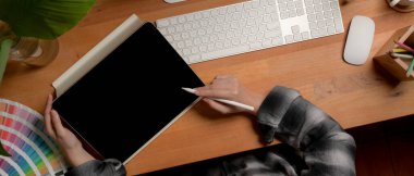 Ahşap ofis masasında dijital tablet, bilgisayar, malzeme ve dekorasyonla çalışan kadın sanatçının genel görüntüsü.