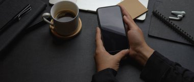 Karanlık modern ofis masasında ofis malzemeleriyle akıllı telefon tutan erkek işçilerin kırpılmış görüntüleri.