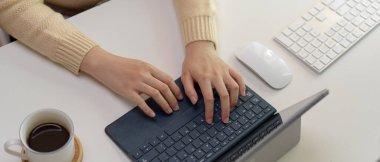 Beyaz ofis masasında, kahve fincanı ve bilgisayar aygıtlarıyla dijital tablet klavyede yazan kadın çalışanın kırpılmış görüntüsü.