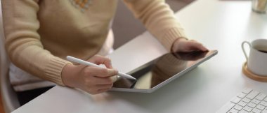 Kadın çalışanın dijital tablete yazı yazışının yan görüntüsü. Ofis odasında bilgisayar ve fincanla beyaz masaya stilus kalemle yazılmış.