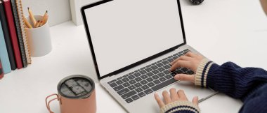 Beyaz masadaki dizüstü bilgisayarda buzlu kahve fincanı, kitaplar ve kırtasiye malzemeleriyle yazı yazan serbest çalışan bir kadın. 