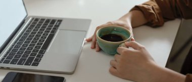 Kadın kahve fincanı tutarken beyaz masadaki akıllı telefonlu dizüstü bilgisayara bakarken yan görünüm