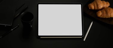 Kahve, kruvasan ve kırtasiye malzemeleriyle birlikte koyu ofis masasının üzerindeki boş ekran tabletinin kırpılmış görüntüsü. 