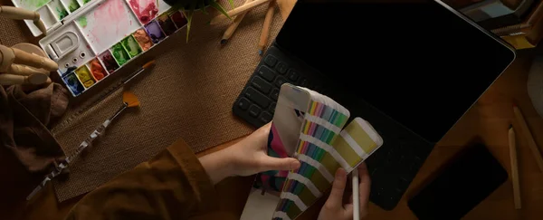 Overhead shot of female designer choosing colour on colour swatch while working with tablet on wooden table