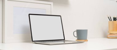 Laptop, kahve fincanı, beyaz masa üzerindeki çerçeve ve kırtasiye malzemeleriyle modern çalışma masasının kırpılmış görüntüsü.