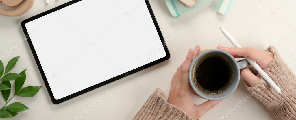 Overhead shot of female student hands holding coffee mug while working with mock up tablet on study table 