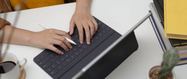 Tablet klavyesinde kahve fincanı, kitap ve dekorasyonla çalışan bir kadının kırpılmış görüntüsü.