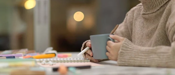 Side view of female student take a break with a cup of coffee while sitting at study table with stationery