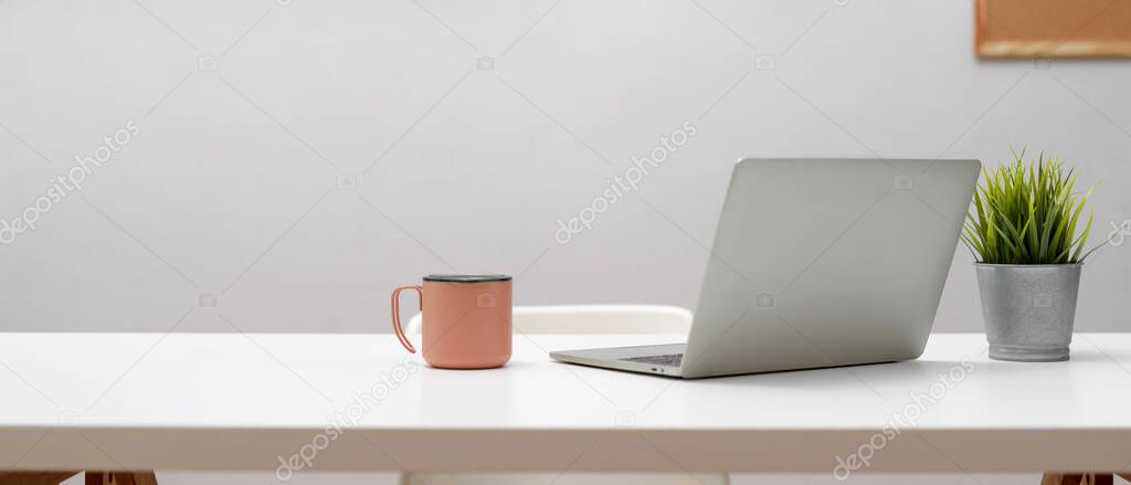 Close up view of home office desk with laptop, mug, plant pot and copy space on white table with white chair 