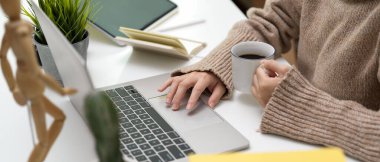Çalışma alanında elinde bir fincan kahve tutarken dizüstü bilgisayar kullanan bir kadının yan görüntüsü