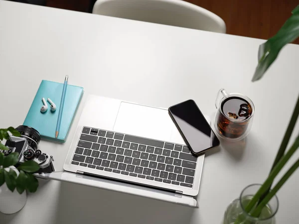 Top view of workspace with laptop, smartphone, camera, supplies and decorations on white table