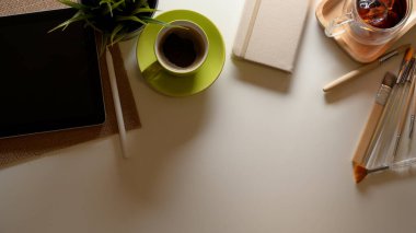 Dijital tableti, kahve fincanı, günlük defteri, boya fırçası, ağaç kabı ve fotokopi alanı olan ev ofis masasının üst görüntüsü 