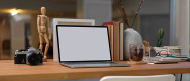 Çalışma alanının boş ekran dizüstü bilgisayarı, kitaplar, malzemeler ve dekorasyonlar oturma odasındaki ahşap masada.