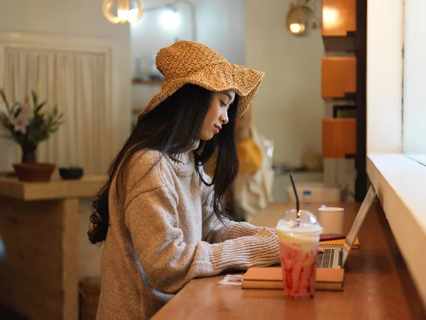 Side view of female in sweater and hat working with laptop and stationery while sitting at counter bar in coffee shop