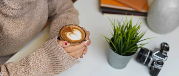 Side view of female in sweater hands holding a cup of coffee on table with camera and supplies