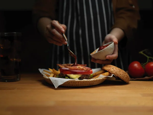 在厨房的木制桌子上 女性厨师在新鲜美味的咸肉汉堡上加番茄酱的近景 — 图库照片