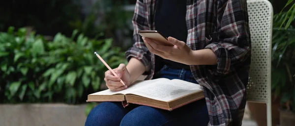 Close up view of female student reading book and using smartphone while sitting in garden