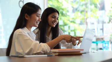 Cam duvar kütüphanesinde dizüstü bilgisayar ve kırtasiyeyle beyin fırtınası yapan kız öğrencilerin portresi 