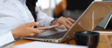 Çalışır durumda dizüstü bilgisayarla çalışırken akıllı telefon kullanan kadın işçinin yan görünümü 