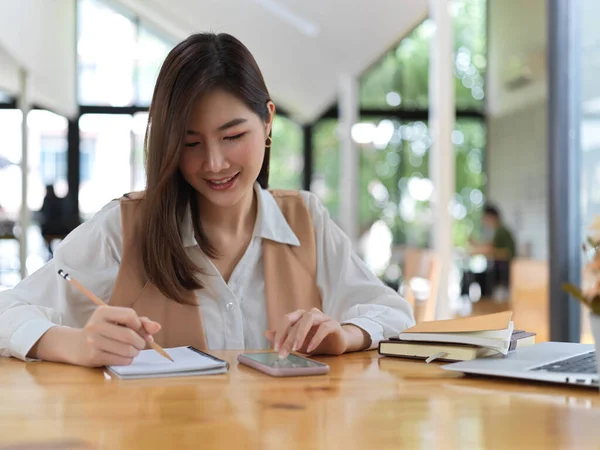Portrait of female college student searching information on smartphone and write it down in notebook