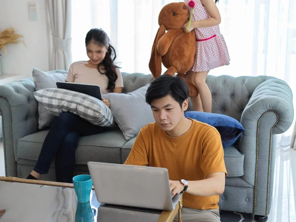 Happy family in living room, mother using digital tablet and father working with laptop while daughter playing with stuffed toy