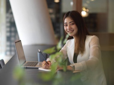 Kırtasiye ve dizüstü bilgisayarla çalışırken gülümseyen ve kameraya bakan kadın ofis çalışanının portresi