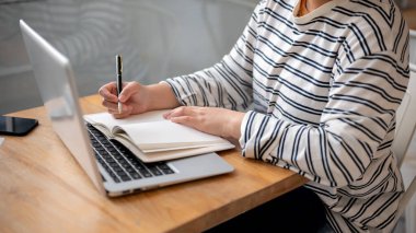 Günlük kıyafetlerle bir kafede çalışan bir kadının dizüstü bilgisayarı üzerinde çalışırken not defterine not aldığı bir fotoğraf..