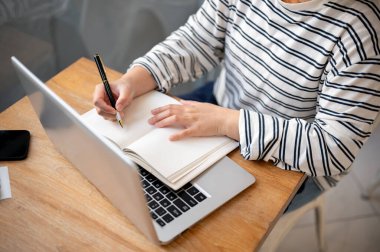 Günlük kıyafetlerle bir kafede çalışan bir kadının dizüstü bilgisayarı üzerinde çalışırken not defterine not aldığı bir fotoğraf..