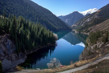 Dağ gölü karlı tepelerin arka planına karşı. Kıyılar boyunca sık bir orman var..