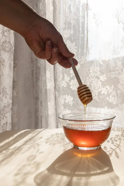 Woman\'s hand holding a honey spoon pouring liquid honey in a glass bowl. Window sunlight on the curtains in the back. Ingredient for desserts, golden light and honey.