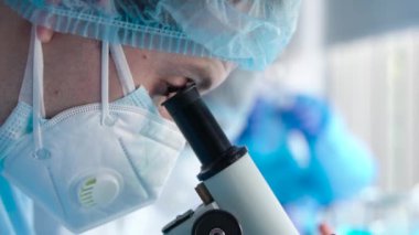 Biyoteknoloji laboratuvarında tıbbi maske takan bilim adamlarının ya da araştırmacıların yavaş çekimde mikroskop merceklerini inceleyerek Coronavirus örneğini incelemeleri.