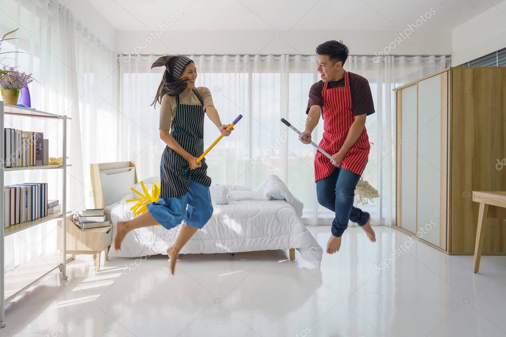 asian young couple having fun doing houseworks together. hasband and wife enjoy housecleaning riding on mop and jump like witches broo